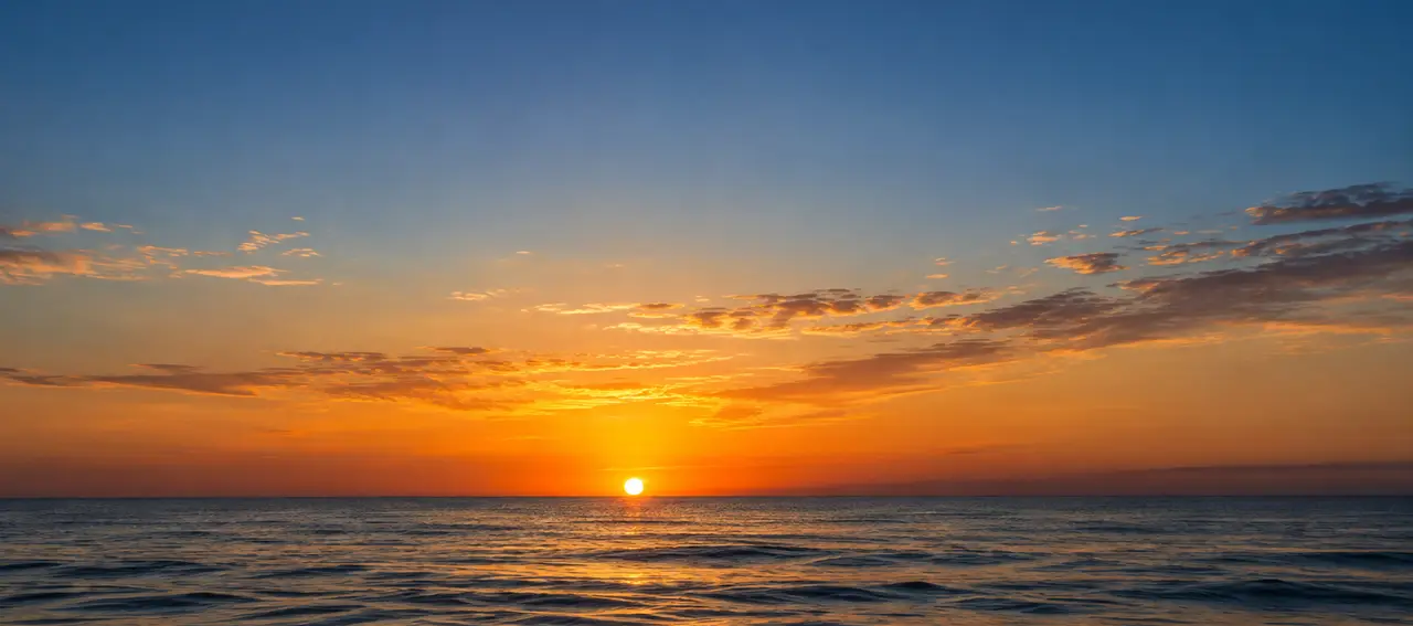The sun just above the sea horizon, warm clouds over calm water
